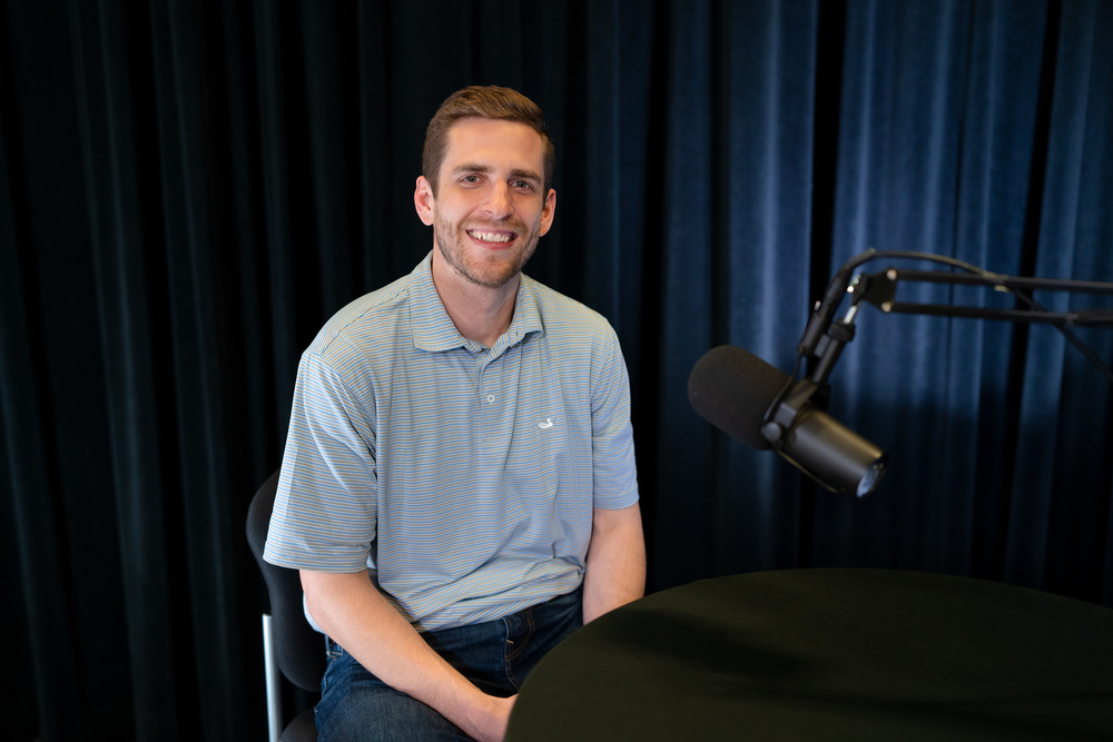 man wearing gray polo shirt smiles at camera as he sits in front of a microphone