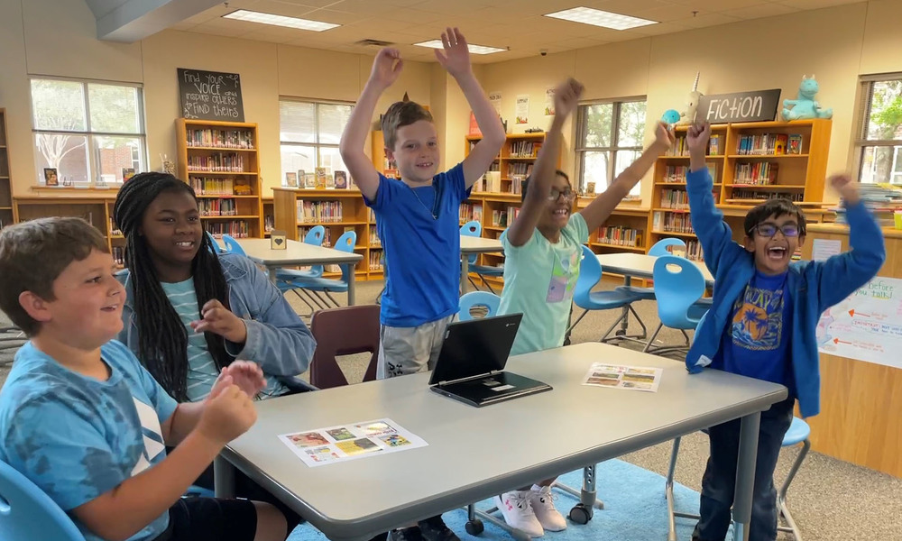 elementary age students around a table in a library. three of them jumping up in celebration