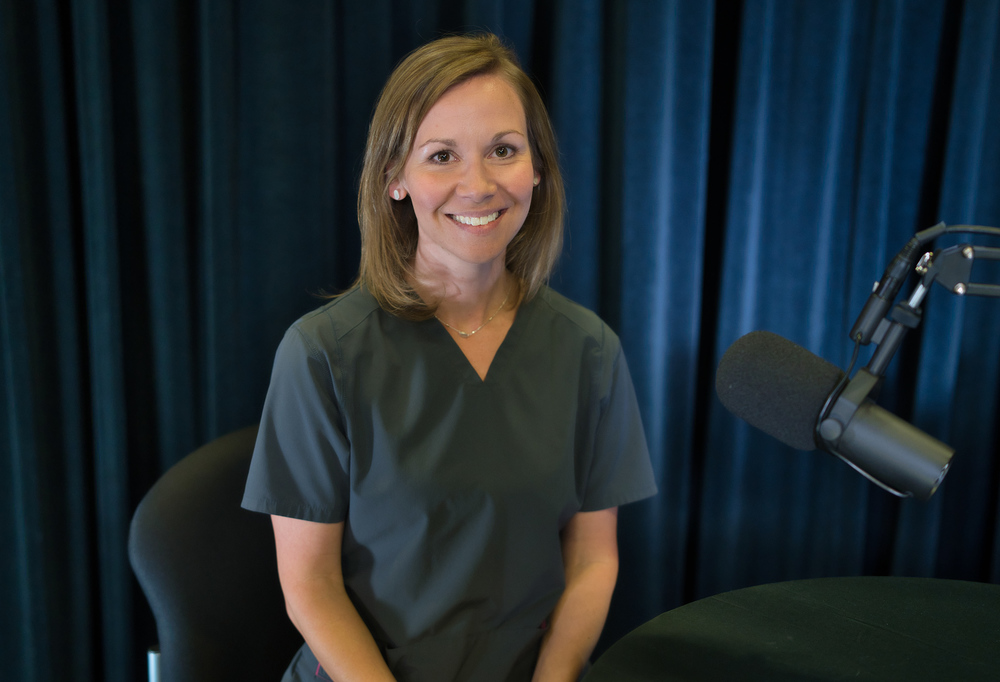 woman wearing gray scrubs sits in front of table with microphone on it smiling at the camera