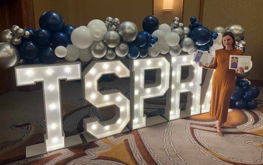 TSPRA sign with lights and blue, silver and white balloons. woman standing next to it holding three certificates