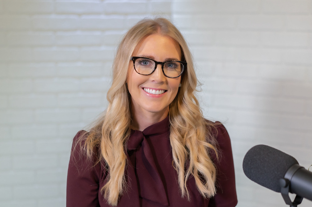 woman with long blond hair, wearing a maroon shirt and black glasses sits behind a microphone