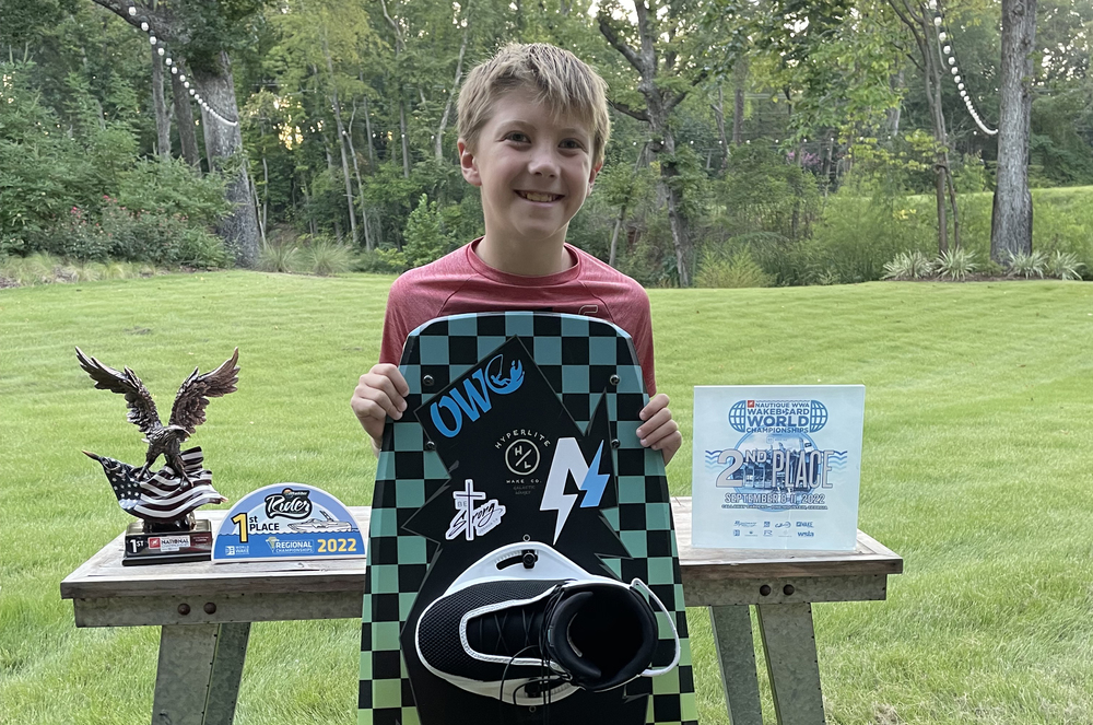 elementary aged boy stands behind his wakeboard with trophies displayed on a table on either side of him