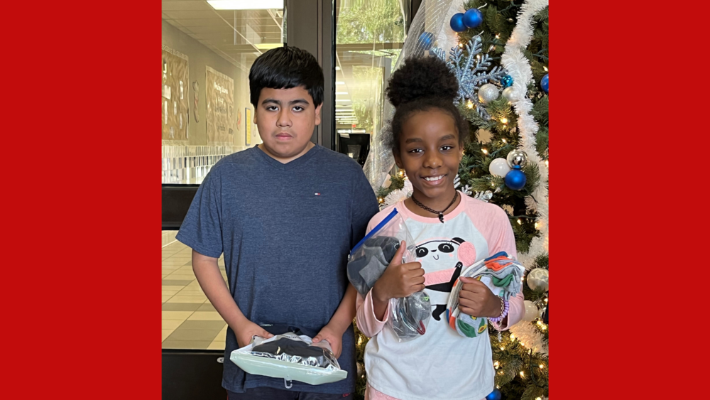 two elementary age students standing in front of a Christmas tree holding socks for a donation