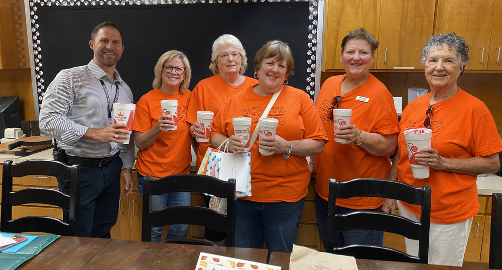 man and five women, all wearing orange t-shirts, standing in a conference room holding smoothies
