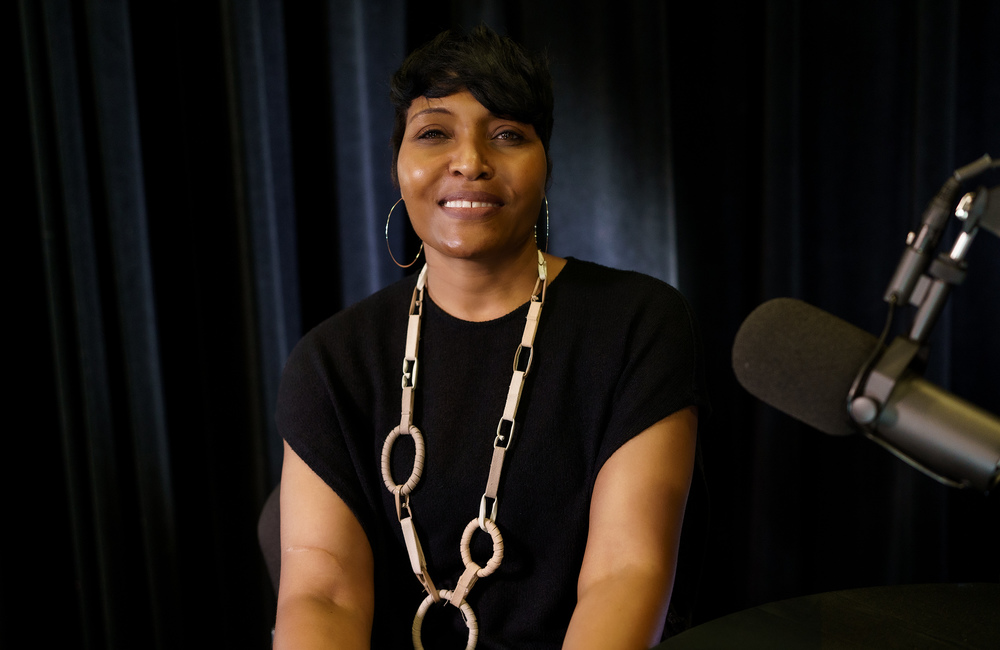 African American woman wearing bulky necklace smiles at the camera with a microphone showing in the foreground