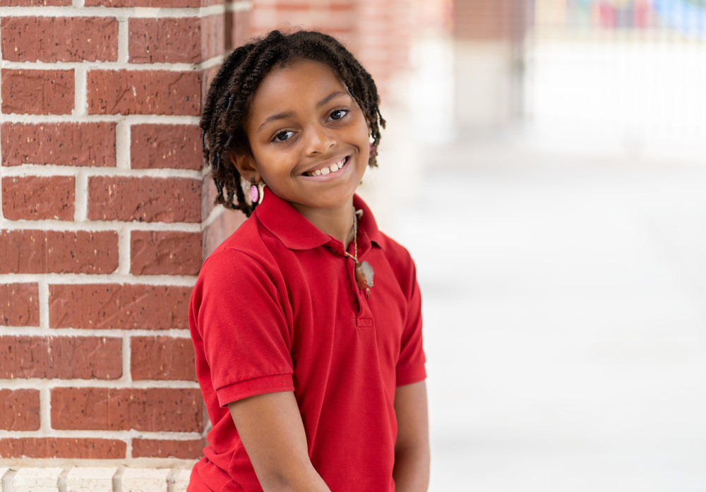 elementary age African-American girl wearing a red polo shirt leaning against a brick wall