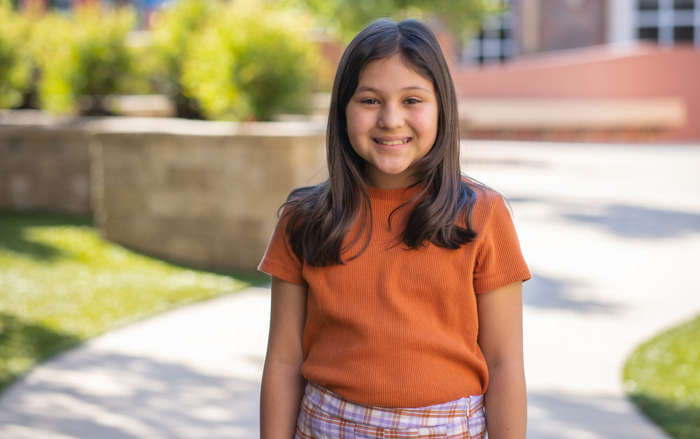 elementary age girl with dark hair wearing an orange shirt and plaid skirt