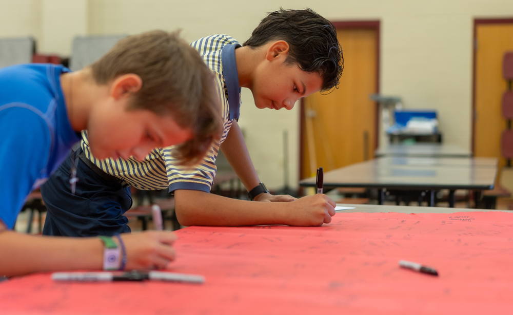 two elementary age boys lean over a table covered with red paper. They are using Sharpies to write their future profession on the paper
