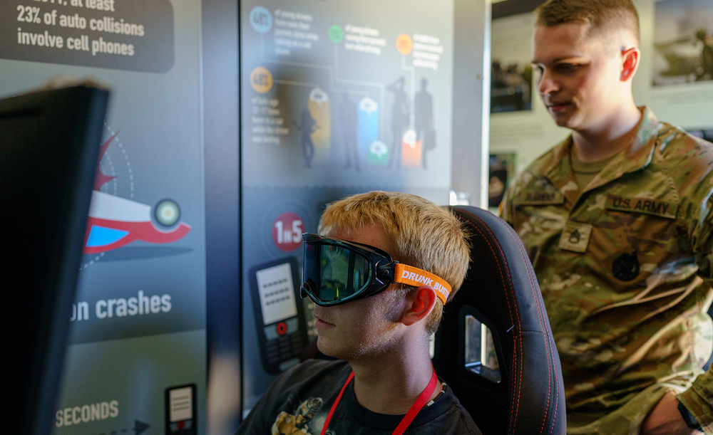 male student sits with goggles on at a virtual reality simulator with male Army serviceman in camo fatigues  looking over his shoulder