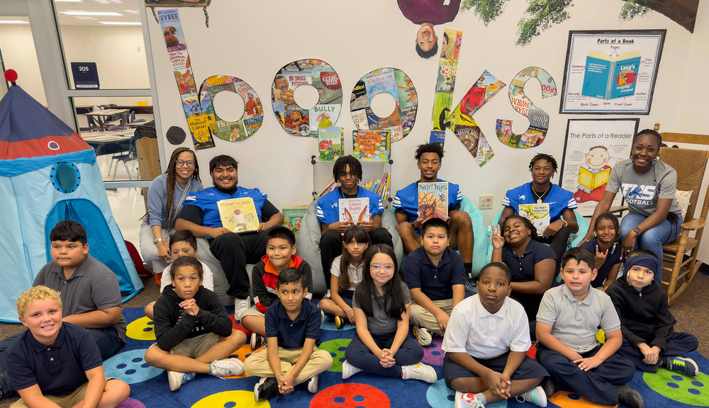 football players sit behind elementary students on the floor in the library