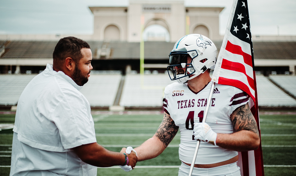 African American man shakes hands with Caucasian man wearing white Texas State football uniform, including helmet. Football player is holding an American flag over his shoulder