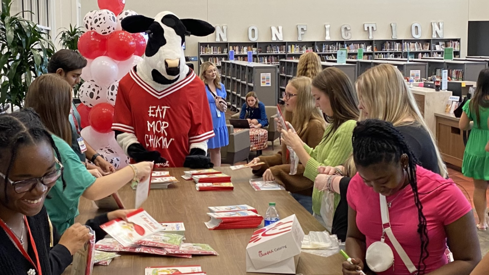 Students packing books in library with Chick-fil-A cow mascot.