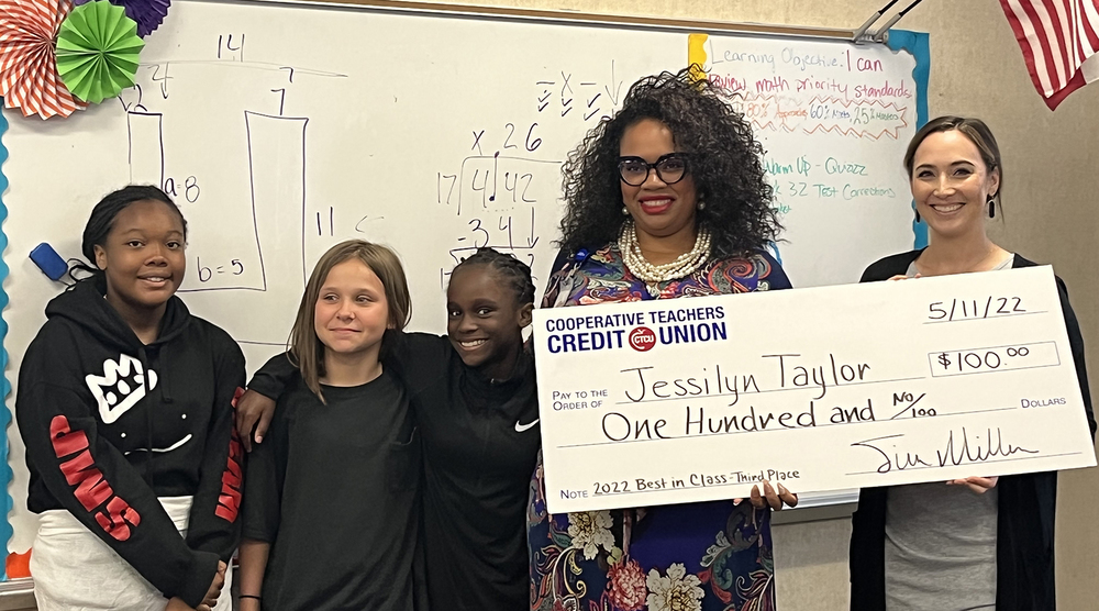 elementary age students stand next to their teacher who is holding a giant check for $100