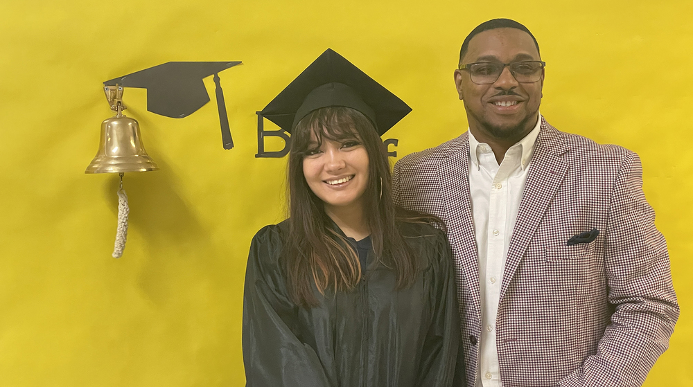girl with long dark hair wearing graduation cap and gown stands next to African American man in tan suit