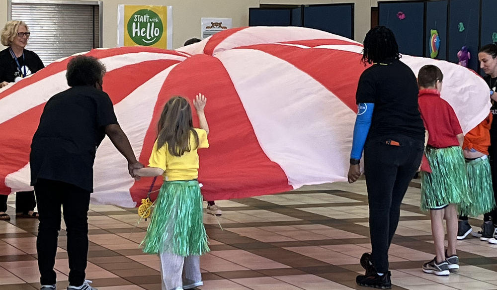 students and adults wearing grass skirts holding red and white parachute with their hands 