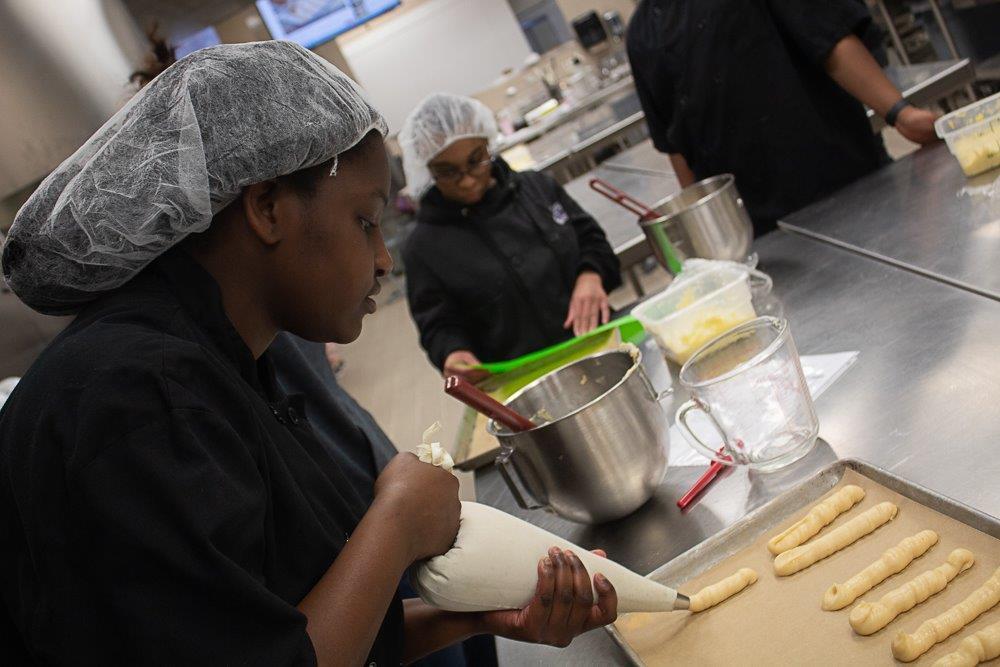 African American student in black jacket wearing a hairnet pipes filling onto a pan. mixing bowls in the background