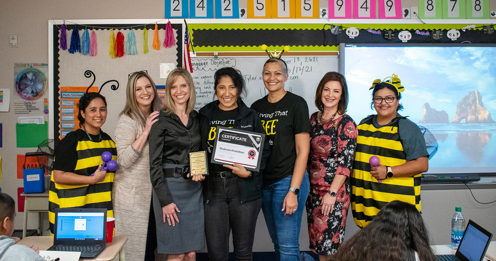 staff members stand around Ms. Khalatbari, who is holding her teacher of the month certificate. Two staff members are wearing "bee" costumes