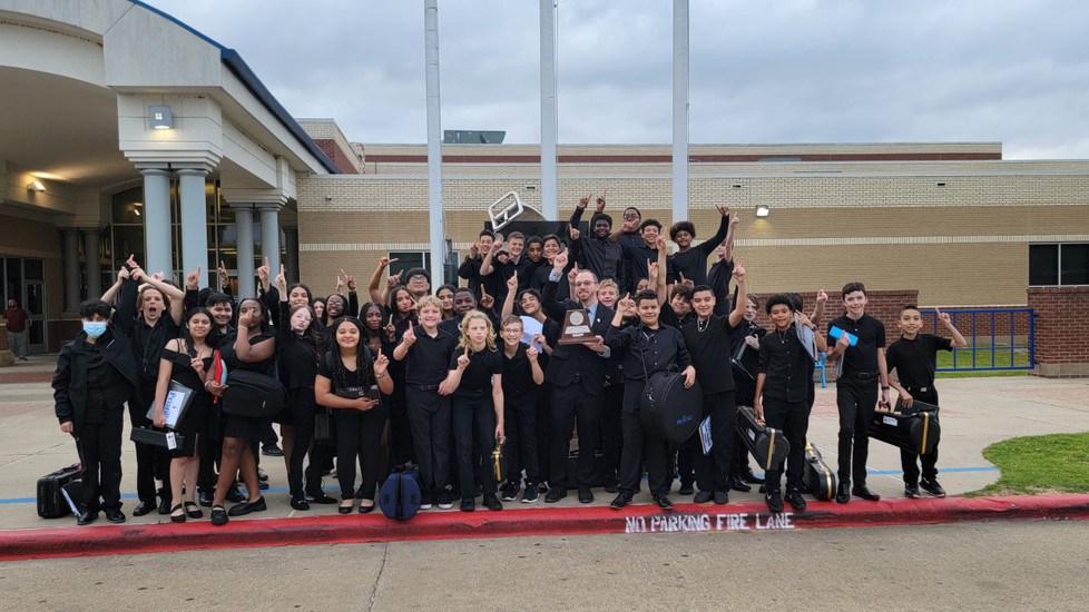 group of middle school students dressed in black shirts and pants stand  in front of their school building