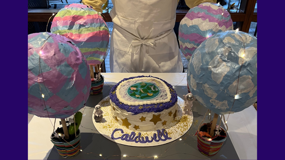 cake decorated with purple trim, white frosting and Caldwell written on the tray holding the cake. four hot air balloon decorations are around the cake