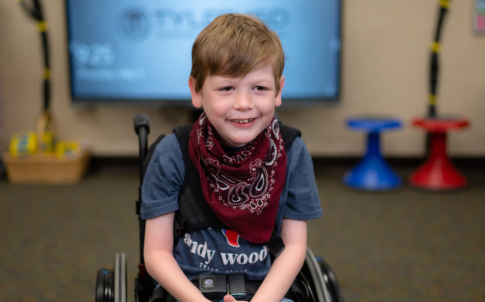 elementary age boy sits in wheelchair wearing gray t-shirt and bandana around his neck