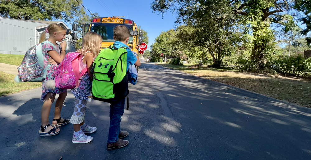 three elementary age children wearing backpacks walking in front of a school bus across the road