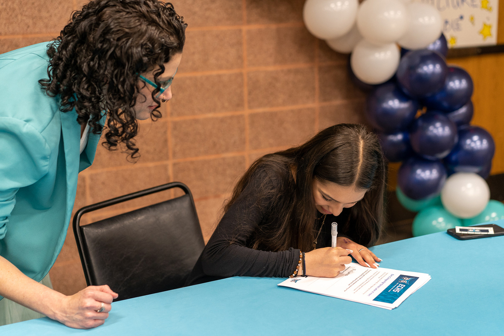 woman with dark curly hair stands over high school student sitting at a table signing a piece of paper