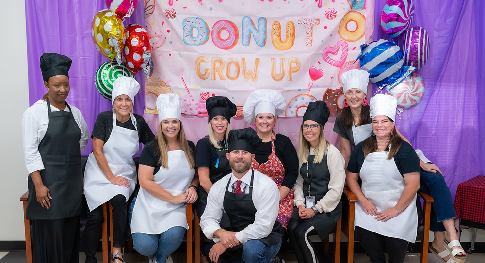 Rice Elementary staff dressed as chefs, wearing aprons and chef hats, sitting in front of a sign that says, "Donut Grow Up"