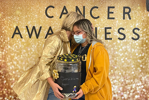 Cancer Awareness. Woman with blonde hair wearing a gold shimmery jacket hugs female student wearing a gold jacket. they are holding a donation bucket in between them.