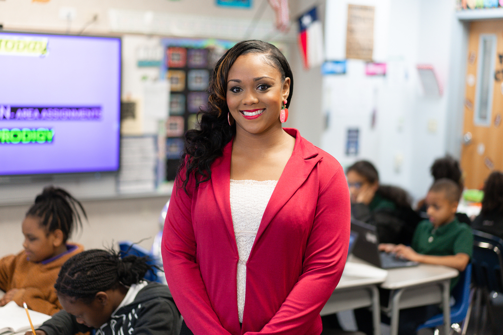 African American woman wearing white shirt with red jacket over it standing in classroom with students at their desks in the background