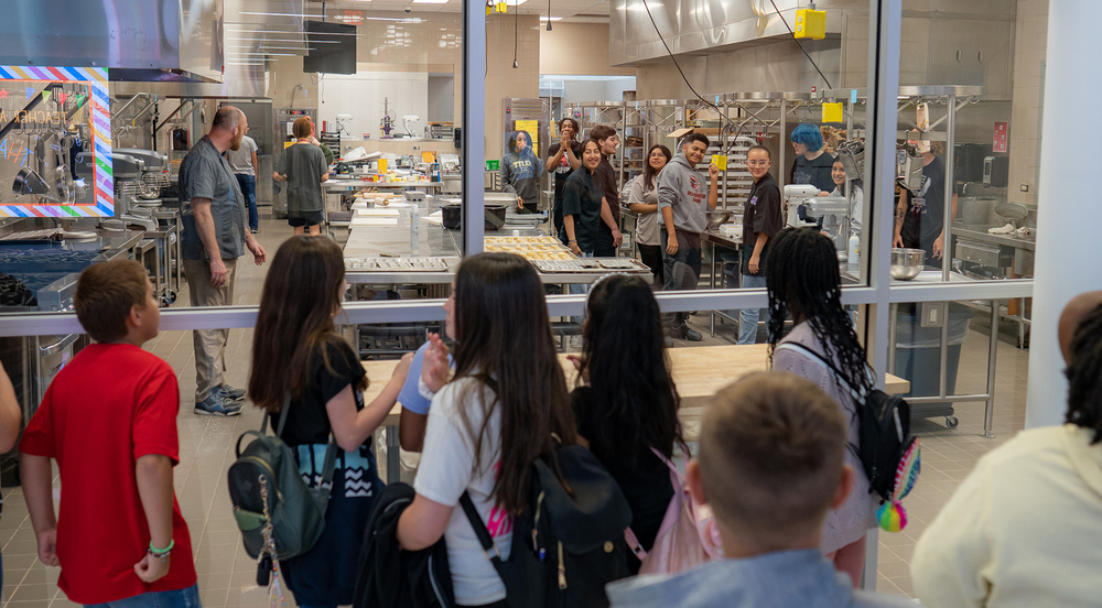 elementary age students look through a glass window at high school students work in culinary kitchen