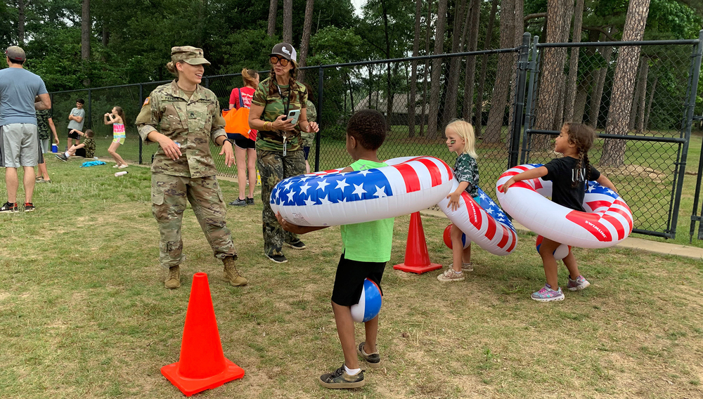 two females wearing Army camo stand in front of small elementary children that have swimming rings around their waists. They are waiting to start an obstacle course