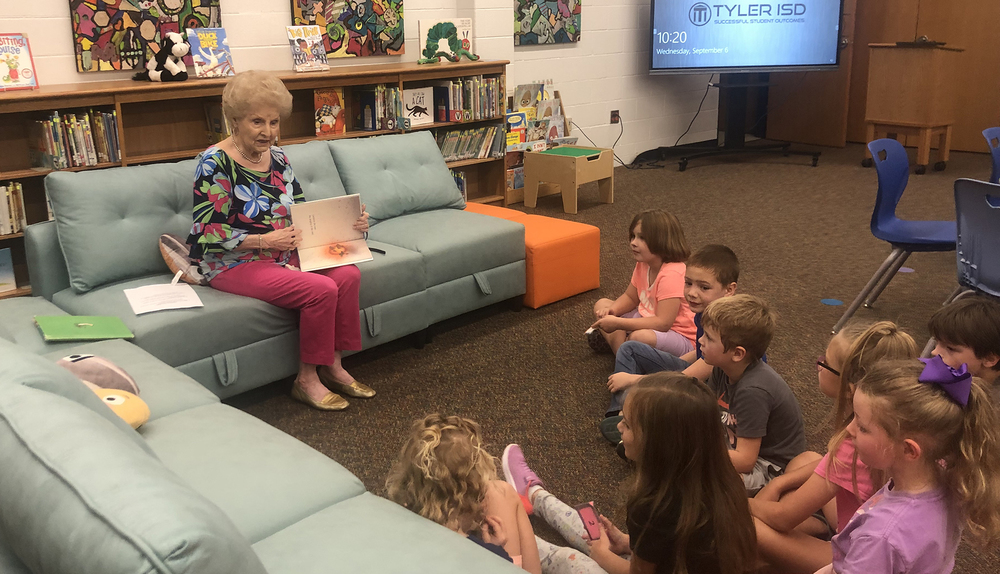 elderly woman sits on couch reading a book to children sitting on the floor