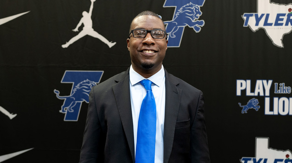 African American man wearing black suit and blue tie standing in front of Tyler Lions Football sign