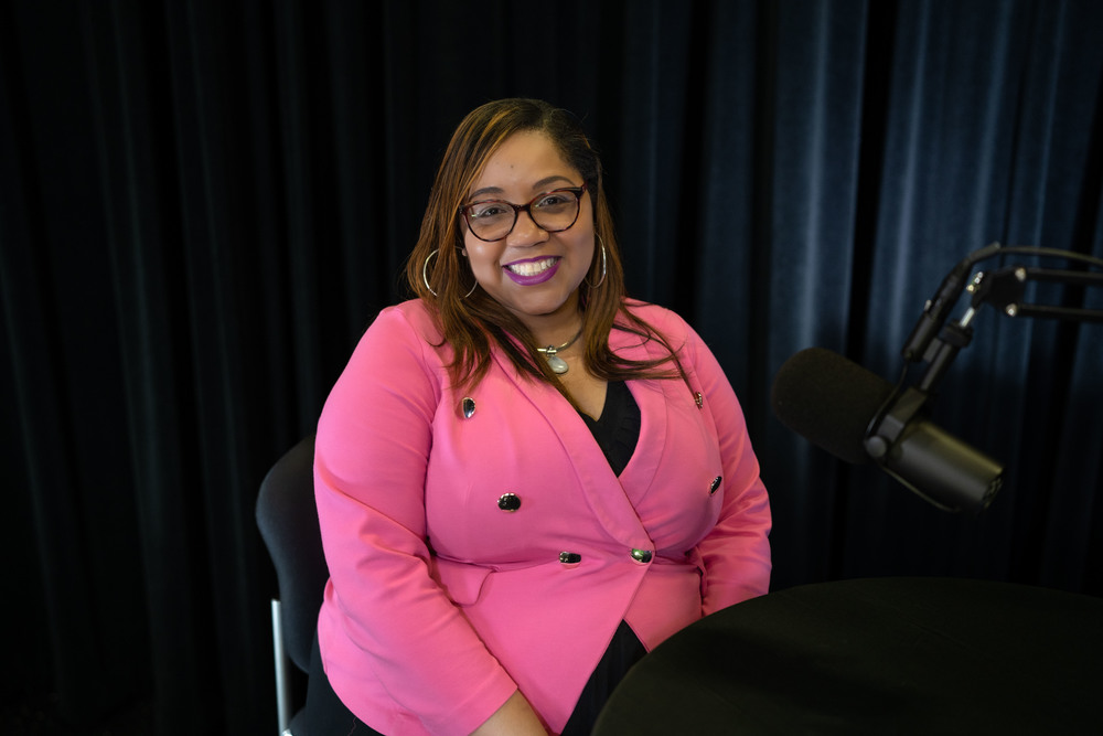 African American woman wearing hot pink jacket smiling at the camera. She is sitting on a chair in front of a microphone