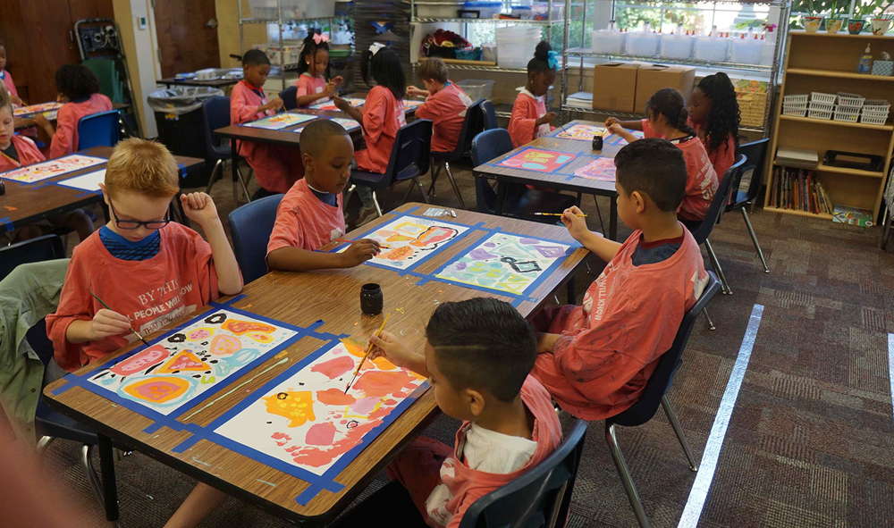 elementary age students wearing orange shirts sitting at desks painting pictures