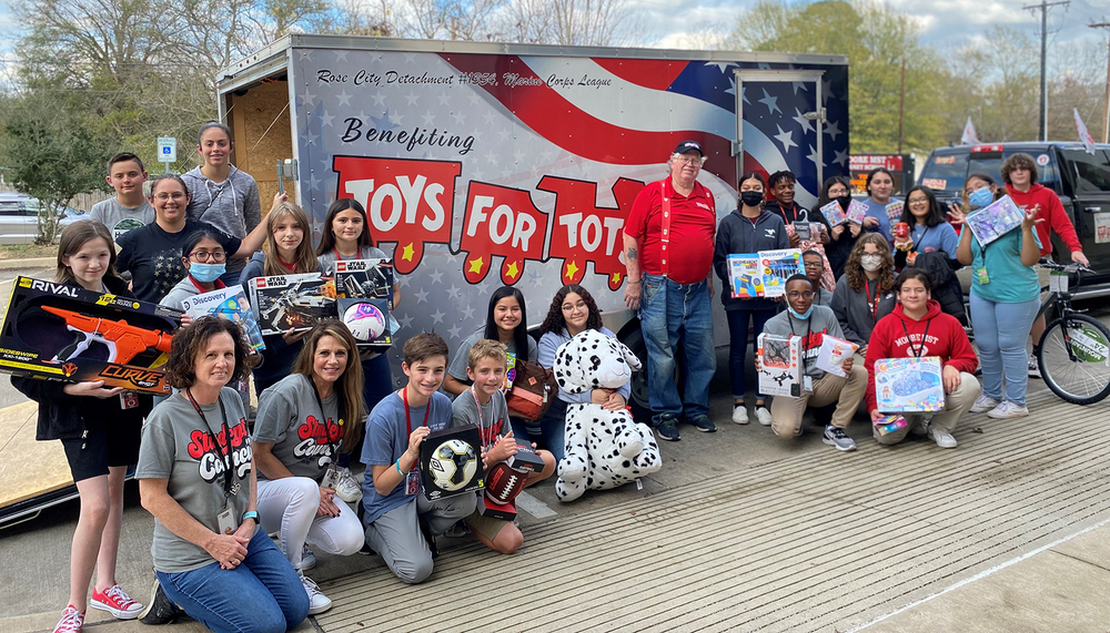 middle school students holding toy donations standing in front of a Toys for Tots trailer