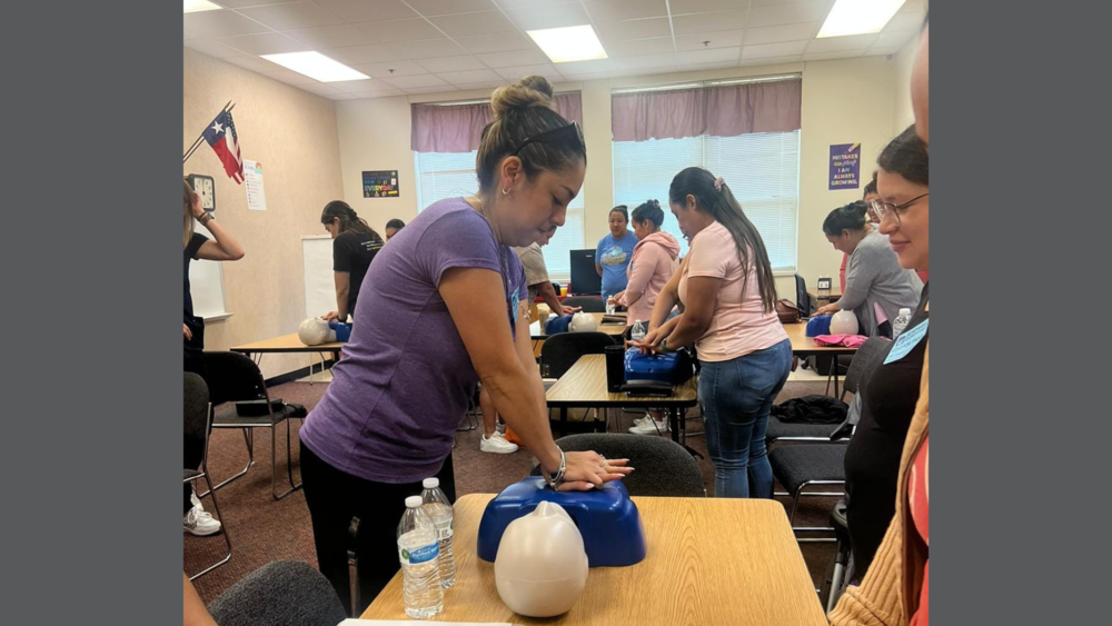 woman wearing a purple shirt and black pants practices chest compressions on a dummy on a table