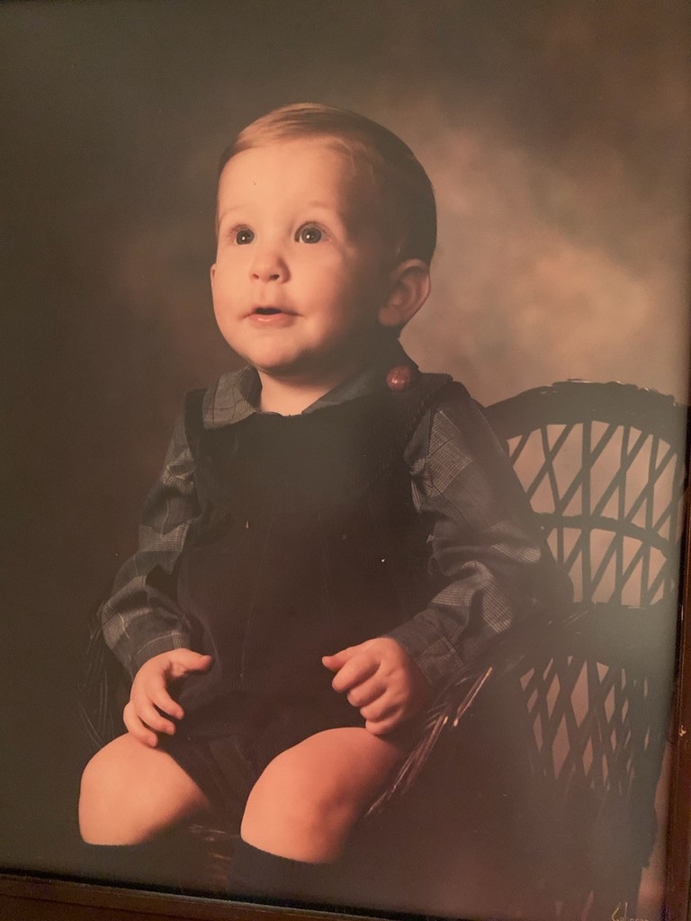 toddler boy sitting in a wicker chair wearing overall shorts, long sleeve shirt and knee high socks. he is looking up into the air with an awestruck expression on his face