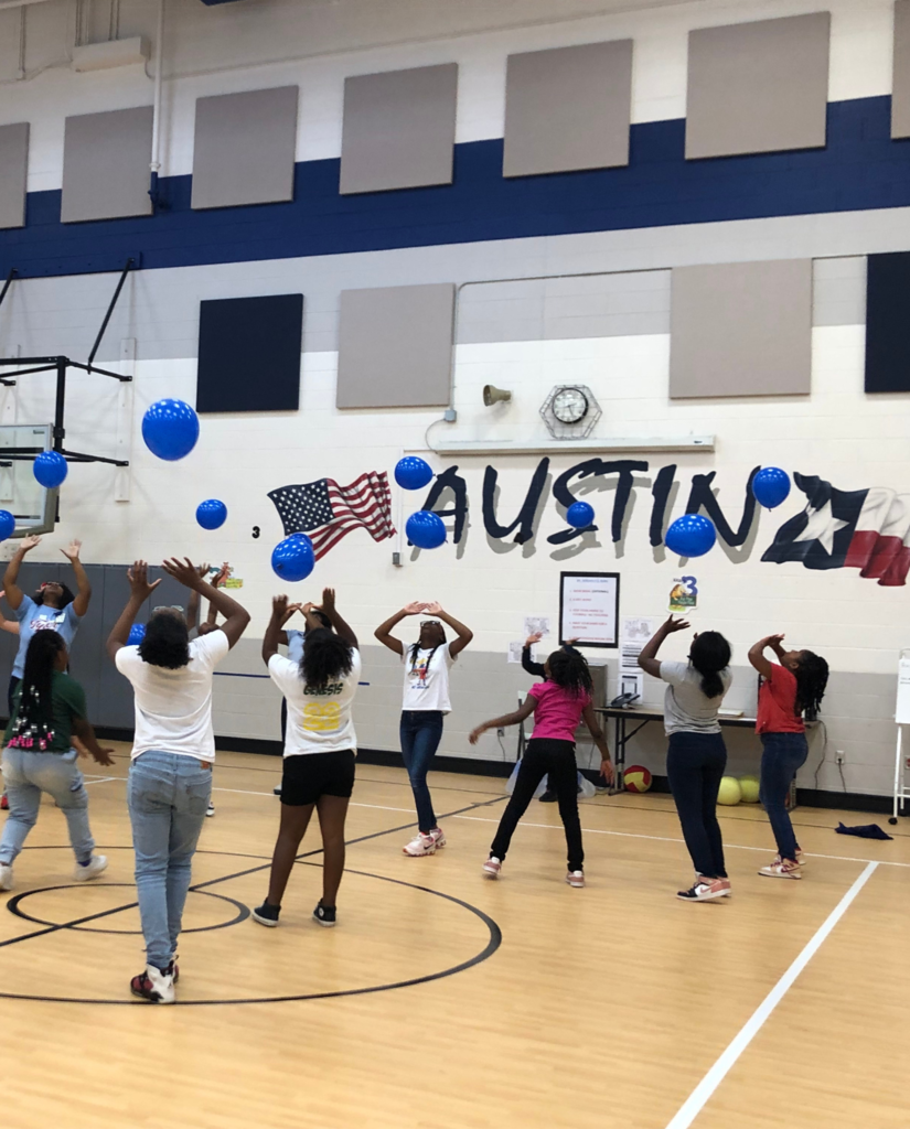 Black female students in white, pink, green and red shirts pushing up a blue balloon in a school gym with the word "Austin" on the wall. 