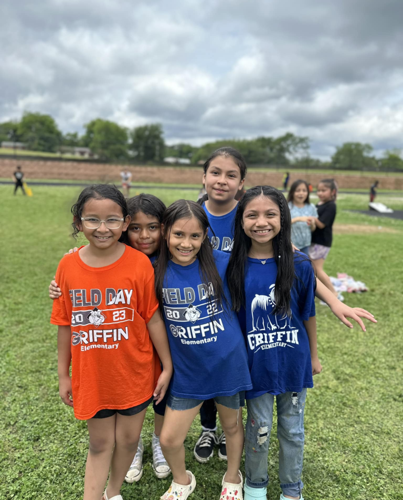 Hispanic females in blue and orange shirts smiling for the camera during an outdoor event. 